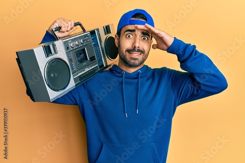 Young hispanic man holding boombox, listening to music stressed and frustrated with hand on head, surprised and angry face