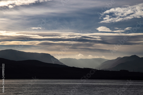 View of Snowdonia mountain range  across the Menai Straits  from Beaumaris. Mountains silhouetted against the late afternoon winter sky  with blue sky  a hint of sunset orange and white fluffy clouds.