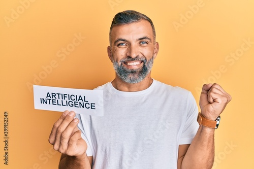 Middle age handsome man holding artificial intelligence word on paper screaming proud, celebrating victory and success very excited with raised arm © Krakenimages.com