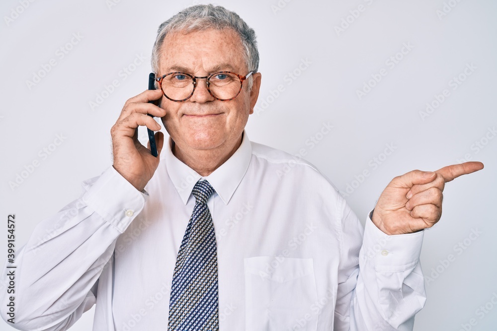 Senior grey-haired man wearing business clothes having conversation talking on the smartphone smiling happy pointing with hand and finger to the side Senior grey-haired man wearing business clothes having conversation talking on the smartphone smiling happy pointing with hand and finger to the side