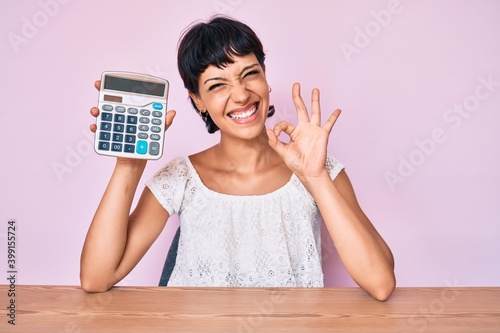 Beautiful brunettte woman showing calculator device doing ok sign with fingers, smiling friendly gesturing excellent symbol