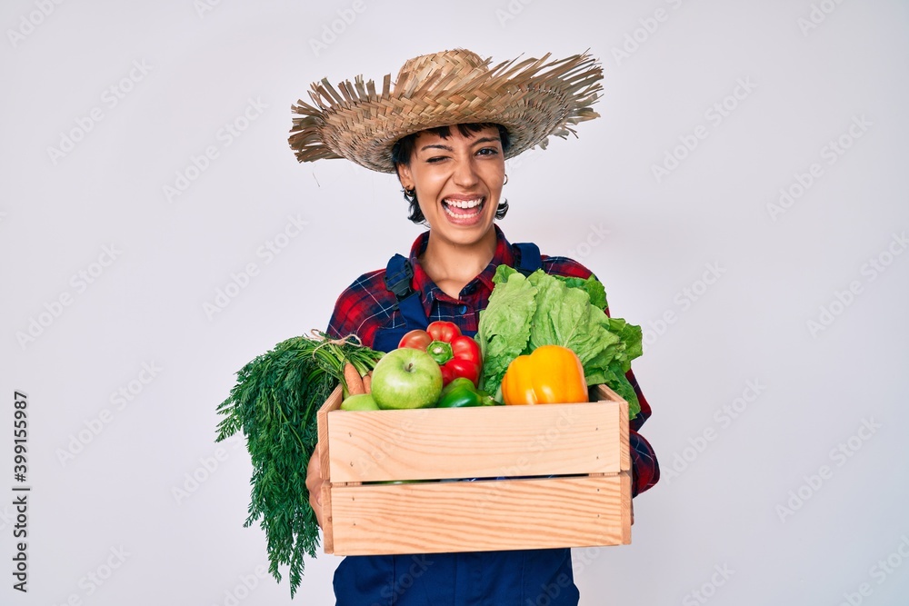 Beautiful brunettte woman wearing farmer clothes holding vegetables winking looking at the camera with sexy expression, cheerful and happy face.