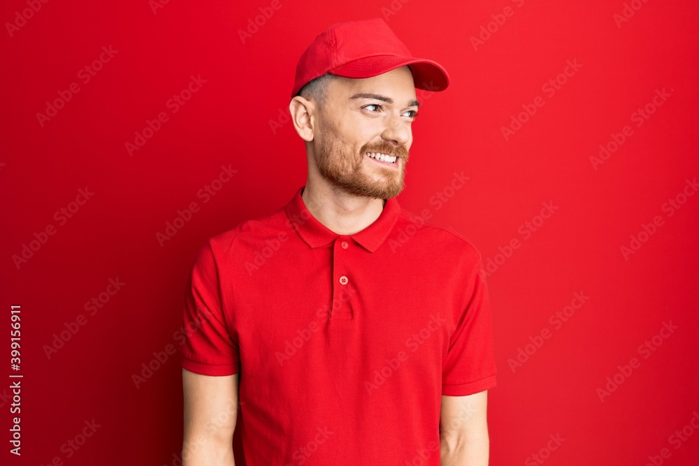 Young redhead man wearing delivery uniform and cap looking away to side with smile on face, natural expression. laughing confident.