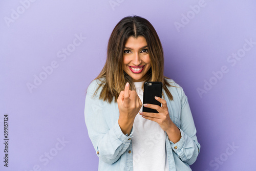 Young indian woman holding a phone isolated pointing with finger at you as if inviting come closer.