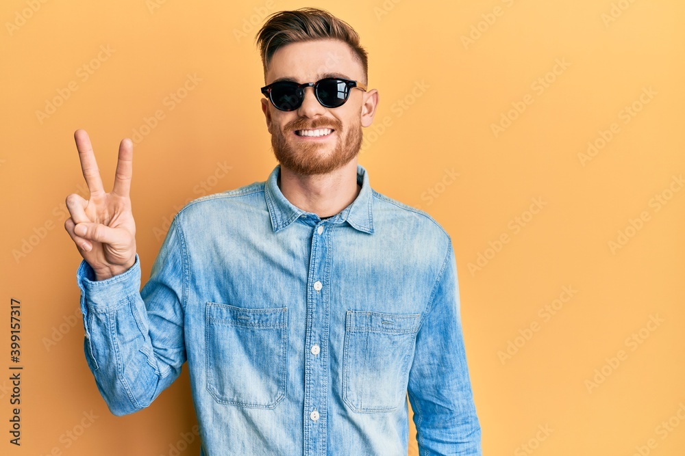 Naklejka premium Young redhead man wearing stylish sunglasses smiling with happy face winking at the camera doing victory sign. number two.
