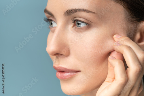 Woman without makeup touching cheeks after glycolic acid peel, has signs of aging skin on her face, close up, isolated on studio blue background