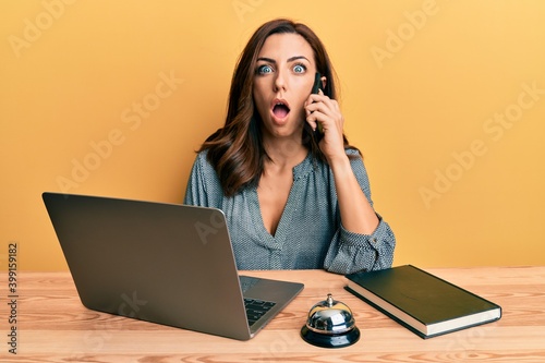 Young brunette woman working at hotel reception talking on the phone afraid and shocked with surprise and amazed expression, fear and excited face.