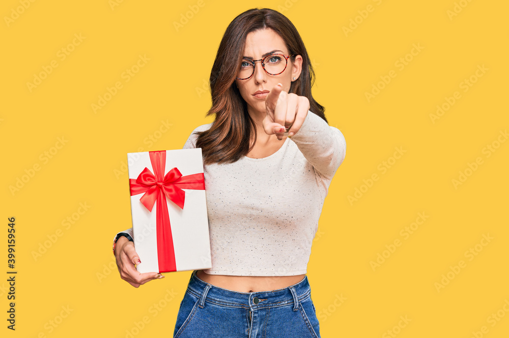 Young brunette woman holding gift pointing with finger to the camera and to you, confident gesture looking serious Young brunette woman holding gift pointing with finger to the camera and to you, confident gesture looking serious