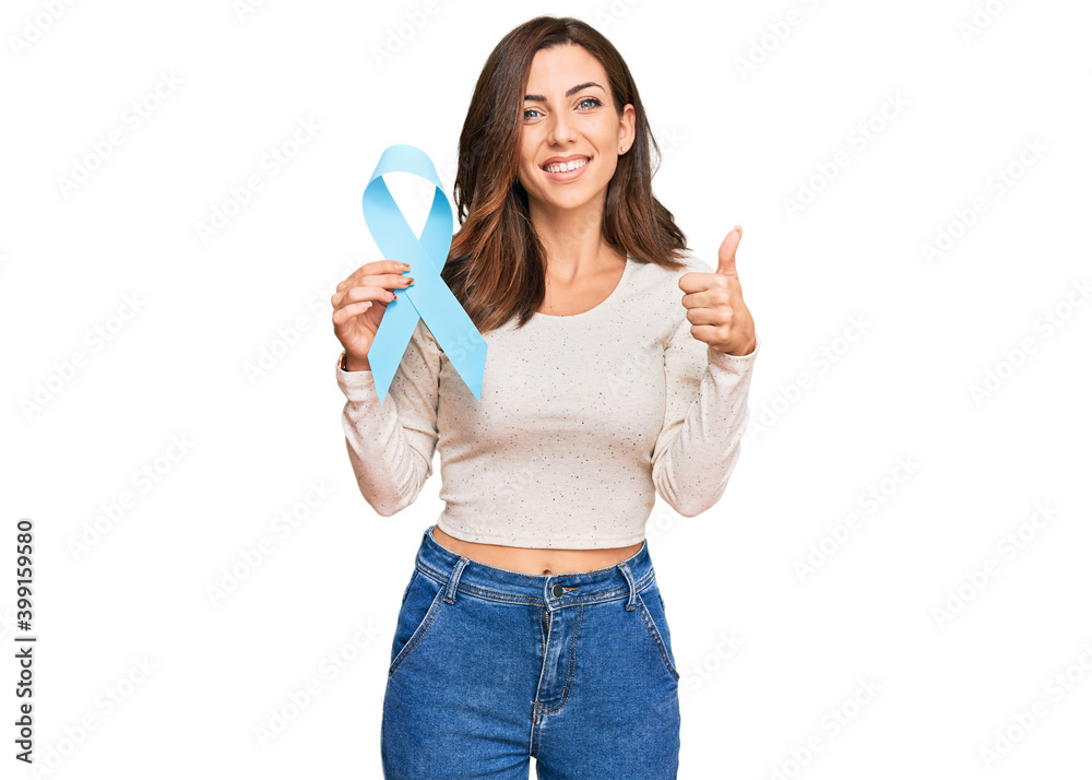 Young brunette woman holding blue ribbon smiling happy and positive, thumb up doing excellent and approval sign Young brunette woman holding blue ribbon smiling happy and positive, thumb up doing excellent and approval sign
