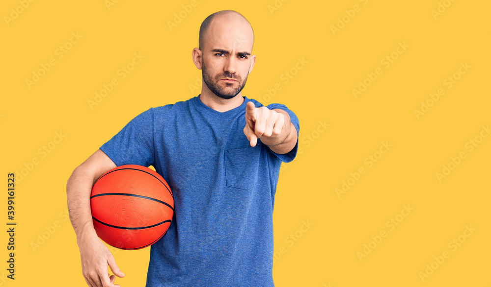 Young handsome man holding basketball ball pointing with finger to the camera and to you, confident gesture looking serious Young handsome man holding basketball ball pointing with finger to the camera and to you, confident gesture looking serious