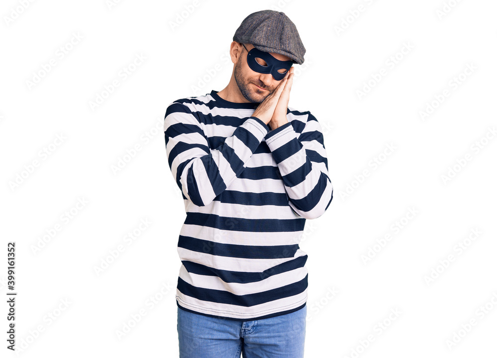 Young handsome man wearing burglar mask sleeping tired dreaming and posing with hands together while smiling with closed eyes. Young handsome man wearing burglar mask sleeping tired dreaming and posing with hands together while smiling with closed eyes.