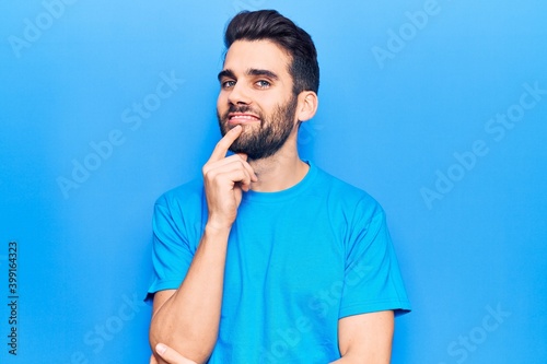 Young handsome man with beard wearing casual t-shirt smiling looking confident at the camera with crossed arms and hand on chin. thinking positive.