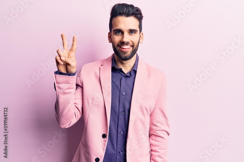 Young handsome man with beard wearing elegant jacket showing and pointing up with fingers number two while smiling confident and happy.