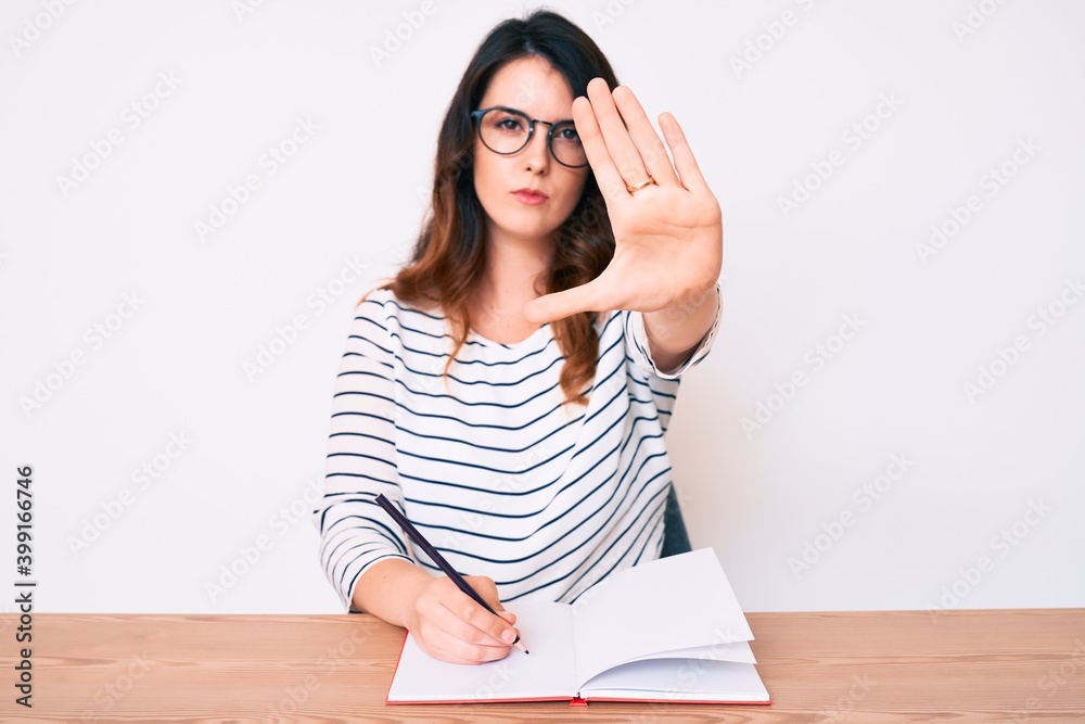 Young beautiful brunette woman writing book sitting on the table with open hand doing stop sign with serious and confident expression, defense gesture Young beautiful brunette woman writing book sitting on the table with open hand doing stop sign with serious and confident expression, defense gesture