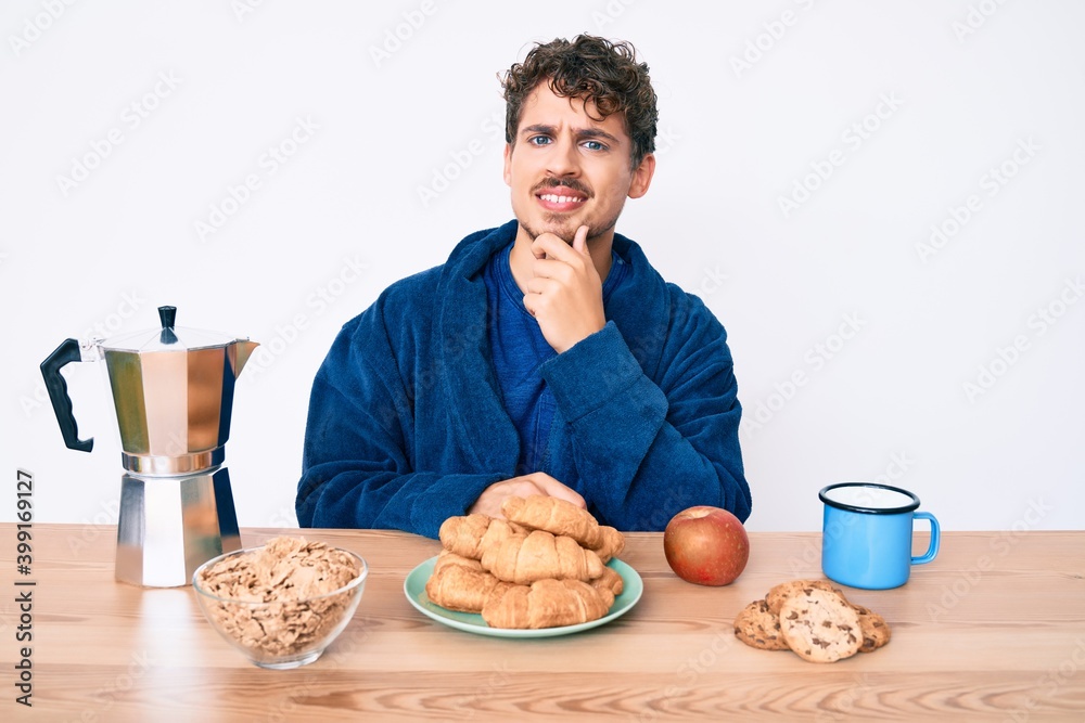 Young caucasian man with curly hair sitting on the table having breakfast smiling looking confident at the camera with crossed arms and hand on chin. thinking positive. Young caucasian man with curly hair sitting on the table having breakfast smiling looking confident at the camera with crossed arms and hand on chin. thinking positive.