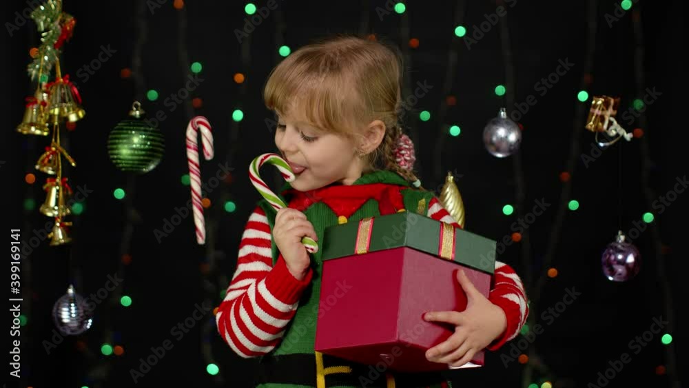 Kid girl in Christmas elf Santa Claus helper costume licking candy cane lollipop on black background with garland. Child with surprise gift box enjoying caramel sweets. New Year holidays celebration