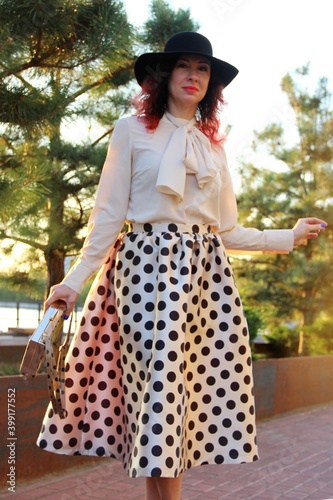 a woman in a black hat, beige blouse and a midi skirt with black polka dots in the bright rays of the sunset walks in a city park on the embankment. portrait of a woman in the sunset. golden hour