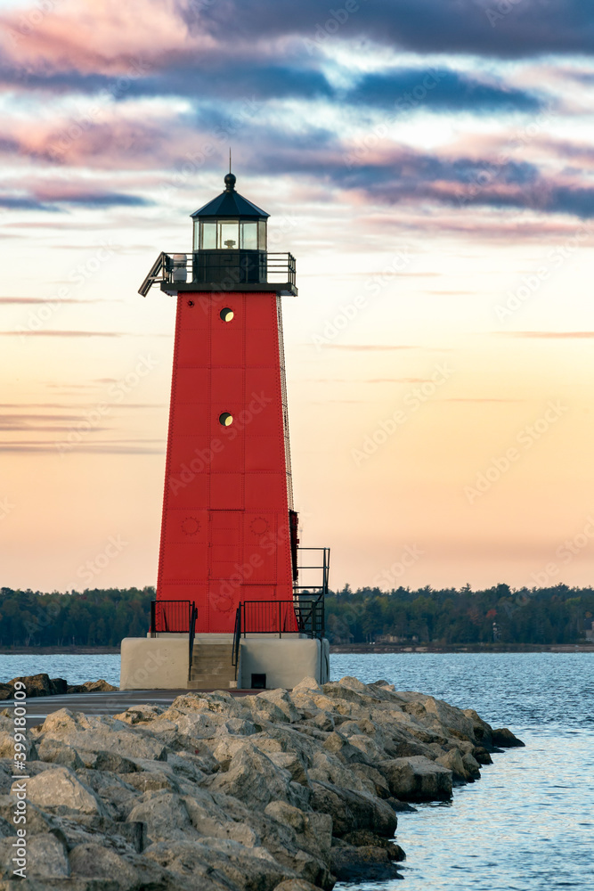 A bright red lighthouse glows in the light of sunrise at Manistique on the Lake Michigan coast of Upper Peninsula Michigan with colorful clouds above.