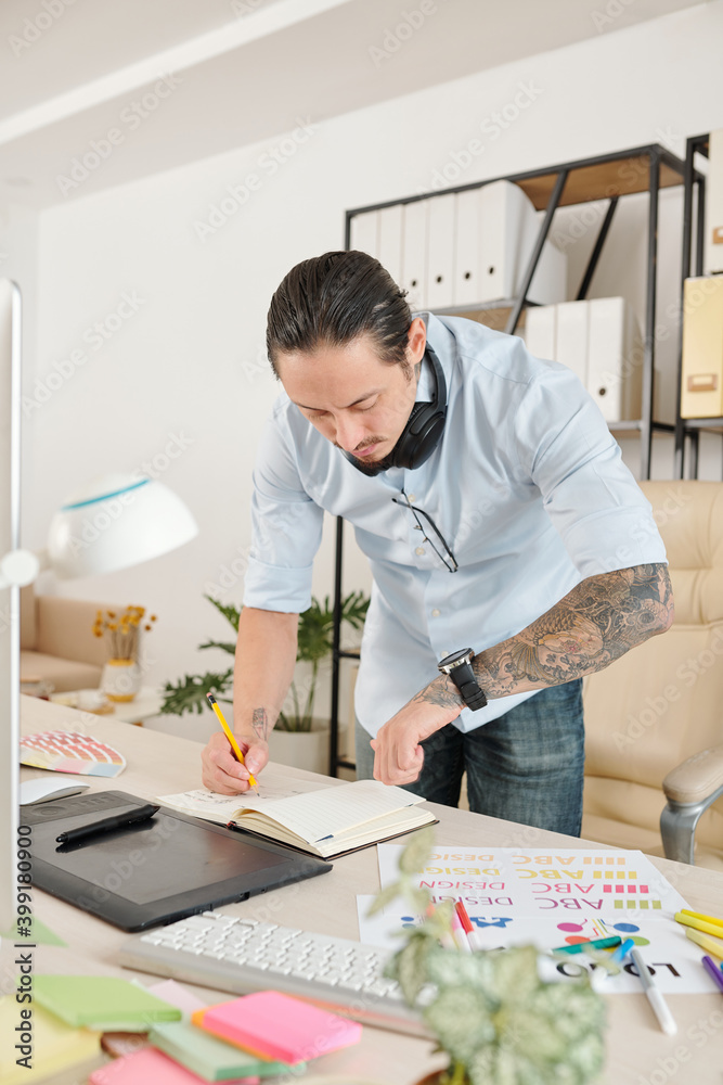Freelance graphic designer checking time on is wrist watch when taking notes in planner Freelance graphic designer checking time on is wrist watch when taking notes in planner