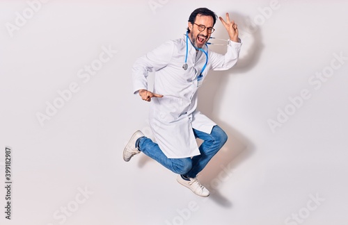 Middle age handsome doctor man wearing stethoscope and coat smiling happy. Jumping with smile on face doing victory sign over isolated white background