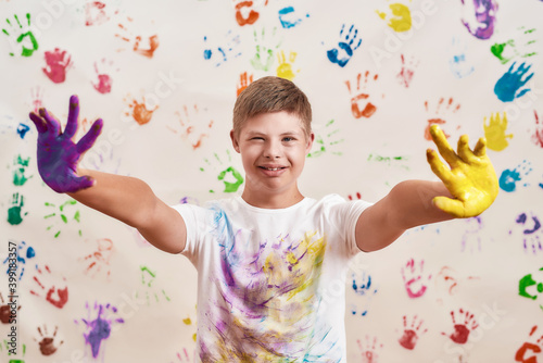 Happy disabled boy with Down syndrome looking at camera while reaching out his hands painted in colorful paints ready for hand prints on the wall