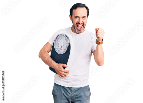 Middle age handsome man holding weighing machine screaming proud, celebrating victory and success very excited with raised arms