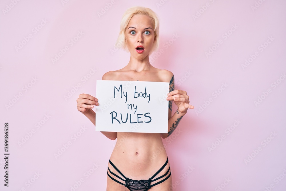 Young blonde woman with tattoo standing shirtless holding banner with my body my rules message afraid and shocked with surprise and amazed expression, fear and excited face. Young blonde woman with tattoo standing shirtless holding banner with my body my rules message afraid and shocked with surprise and amazed expression, fear and excited face.
