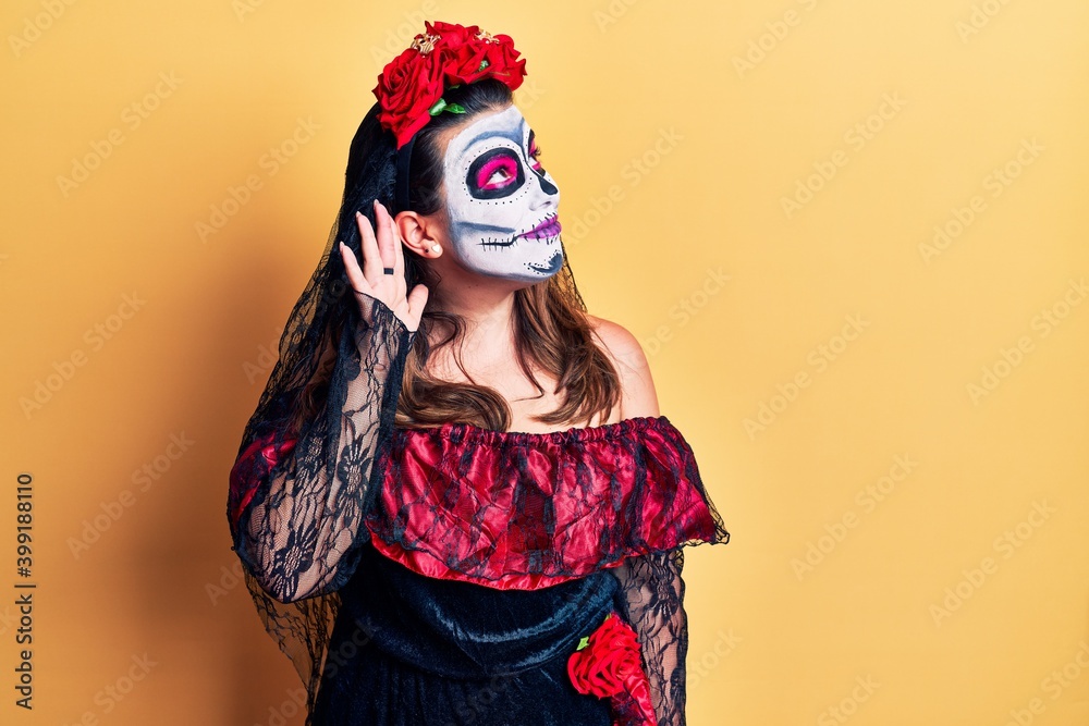 Young woman wearing day of the dead costume over yellow smiling with hand over ear listening an hearing to rumor or gossip. deafness concept.