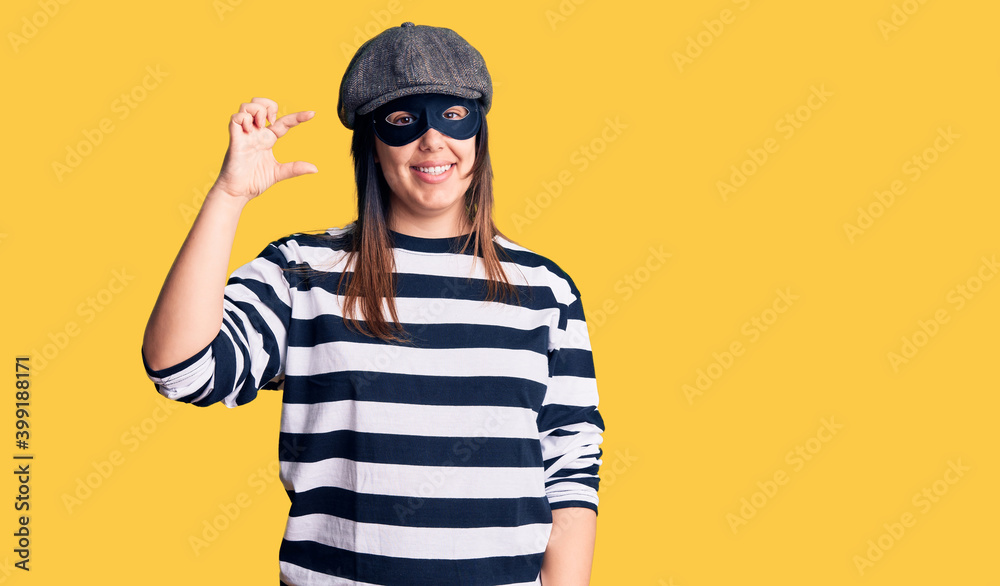 Young beautiful brunette woman wearing burglar mask smiling and confident gesturing with hand doing small size sign with fingers looking and the camera. measure concept. Young beautiful brunette woman wearing burglar mask smiling and confident gesturing with hand doing small size sign with fingers looking and the camera. measure concept.