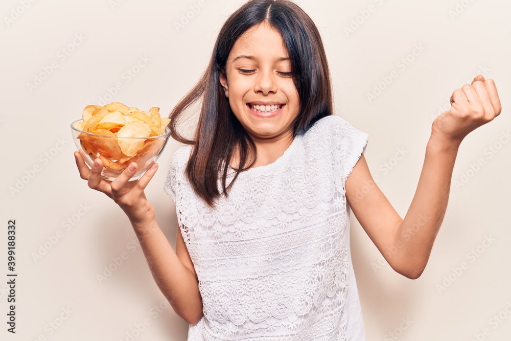 Beautiful child girl holding potato chip screaming proud, celebrating victory and success very excited with raised arms Beautiful child girl holding potato chip screaming proud, celebrating victory and success very excited with raised arms