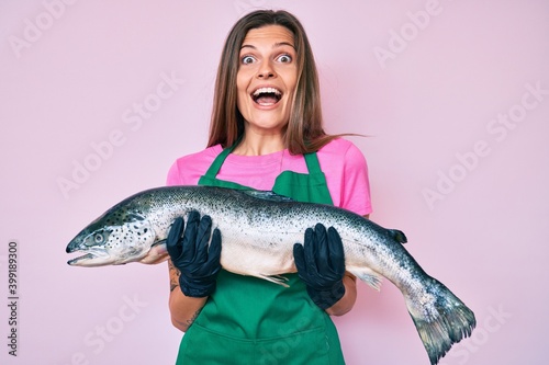 Beautiful caucasian woman fishmonger selling fresh raw salmon celebrating crazy and amazed for success with open eyes screaming excited.