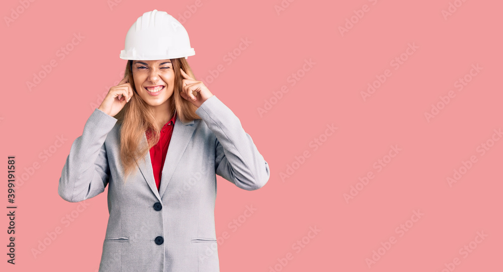Beautiful young woman wearing architect hardhat covering ears with fingers with annoyed expression for the noise of loud music. deaf concept.