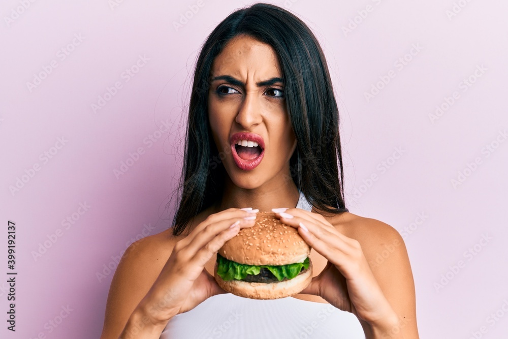 Beautiful hispanic woman eating a tasty classic burger angry and mad screaming frustrated and furious, shouting with anger. rage and aggressive concept.