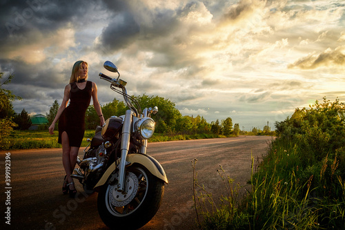Attractive female brunette motorcyclist with motorcycle in a summer evening during sunset. Beautiful girl on nature and sky with sun and clouds background. Adventure concept