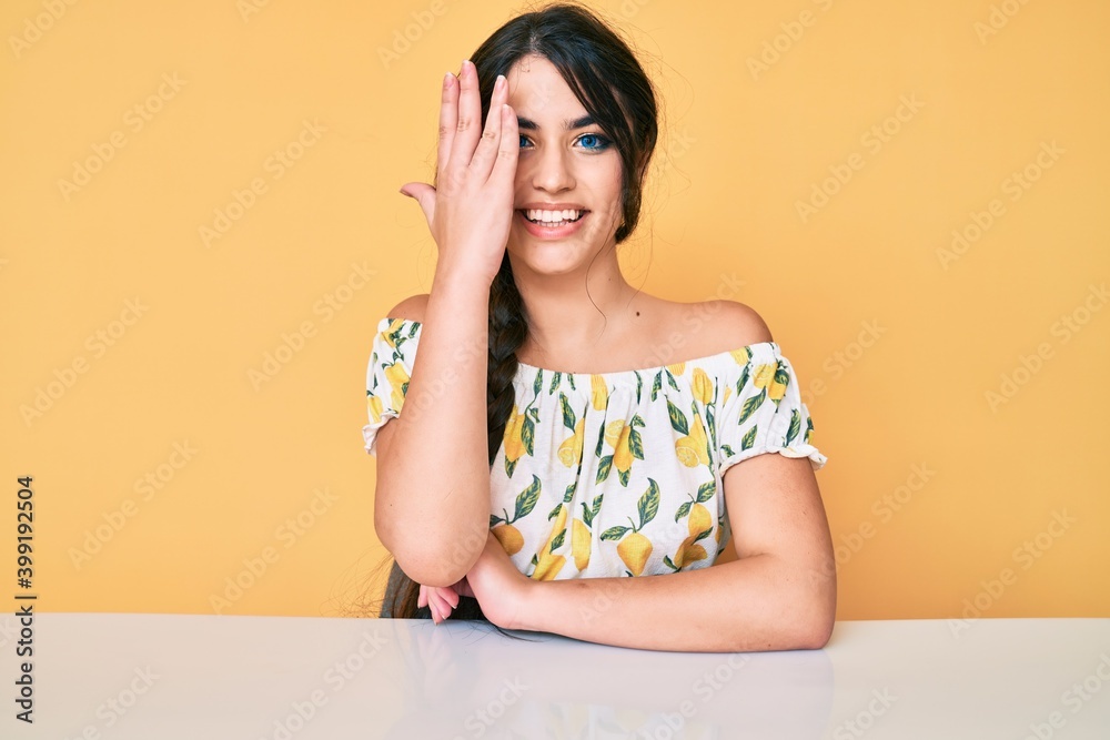 Brunette teenager girl wearing casual clothes sitting on the table covering one eye with hand, confident smile on face and surprise emotion.