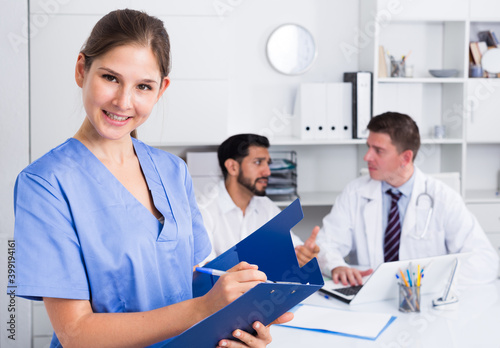 Smiling attractive female doctor writing notes on clipboard in office with colleagues