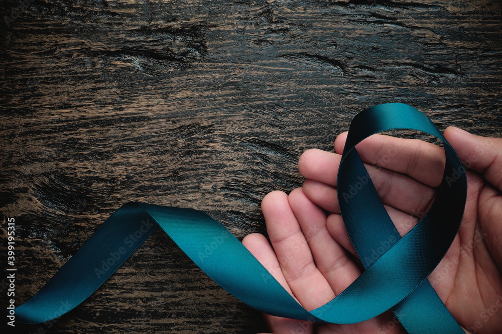 Top view of hand holding teal ribbon on dark background with copy space. Cervical and ovarian cancer, sexual assault, pcos, ptsd, anxiety disorder, agoraphobia and scleroderma awareness concept.