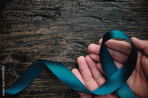 Top view of hand holding teal ribbon on dark background with copy space. Cervical and ovarian cancer, sexual assault, pcos, ptsd, anxiety disorder, agoraphobia and scleroderma awareness concept. photo