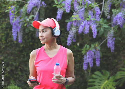 ealthy Asian woman in pink sportswear standing in the garden with purple flowers, hoding bottle of water, mobile phone and wearing headphones. sports concept. photo