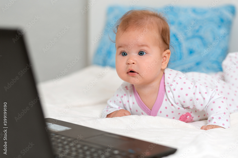 Portrait of amazement baby in sliders lies on the bed and looks at the laptop. The concept of teaching children to modern technologies Portrait of amazement baby in sliders lies on the bed and looks at the laptop. The concept of teaching children to modern technologies