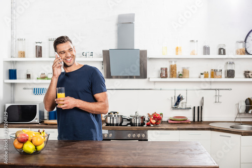  man talking on smartphone while holding glass of orange juice near fruits in kitchen
