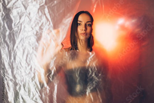 Art portrait. Plastic pollution. Environmental protection. Save nature. Confident woman from future looking through torn wrinkled texture polyethylene film in red neon light.