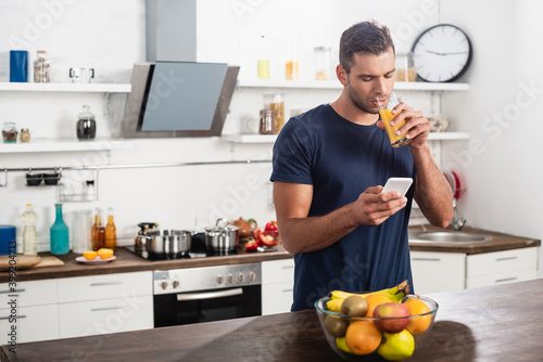 man using smartphone while drinking orange juice near fruits on blurred foreground