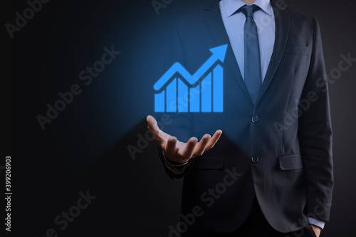 Businessman holding tablet and showing a growing virtual hologram of statistics, graph and chart with arrow up on dark background. Stock market. Business growth, planing and strategy concept.