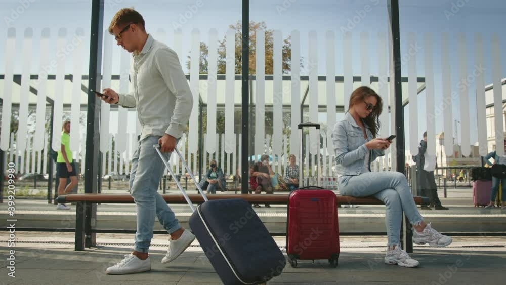 A young woman is sitting at a public transport stop and texting on her smartphone. A man is sitting down next to her and also texting. 4K.