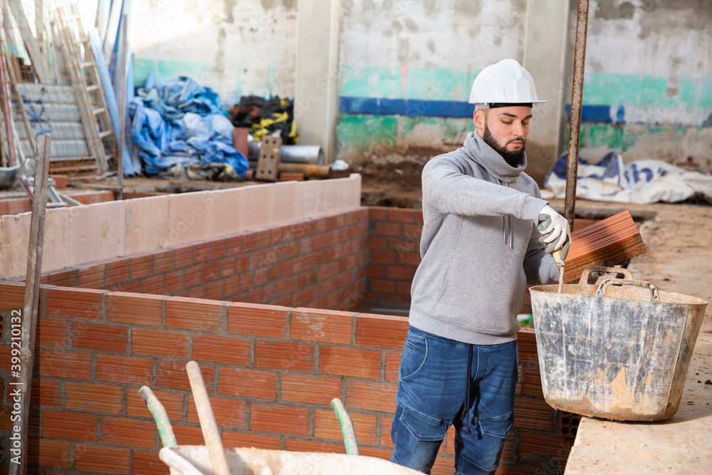 Young bearded bricklayer installing brick wall in building under construction..