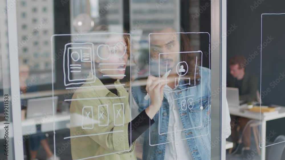 PAN shot of young female UI designer drawing interface ideas on glass wall or window in office and talking to male colleague