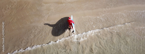Aerial drone ultra wide panoramic photo of Santa Claus coming out of turquoise exotic sandy beach holding SUP or Stand Up Paddle board at Christmas time