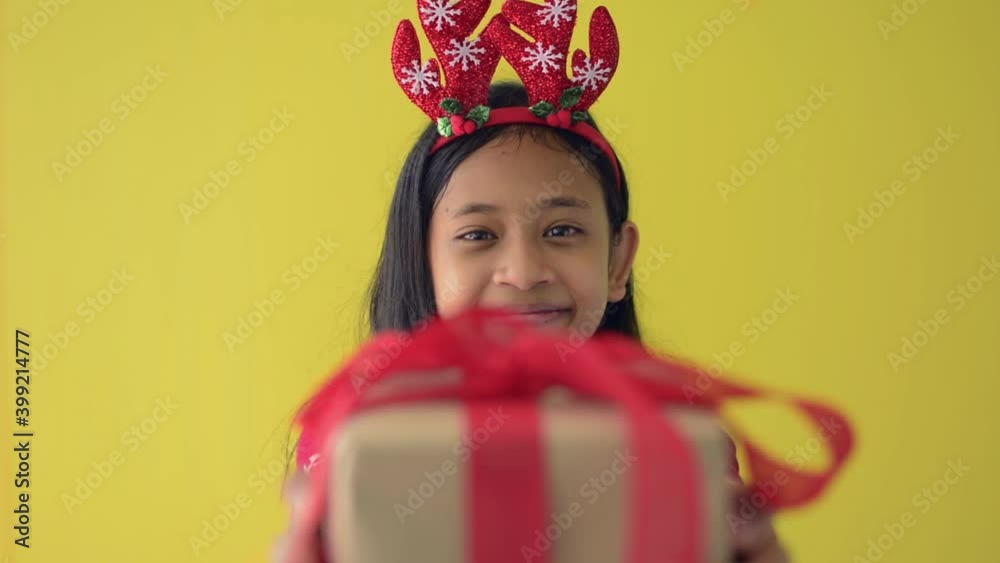 Portrait adorable girl with long hair in red sweater wearing reindeer antlers headband is smiling and giving a gift box while looking at camera isolated on yellow background. Merry Christmas. Portrait adorable girl with long hair in red sweater wearing reindeer antlers headband is smiling and giving a gift box while looking at camera isolated on yellow background. Merry Christmas.