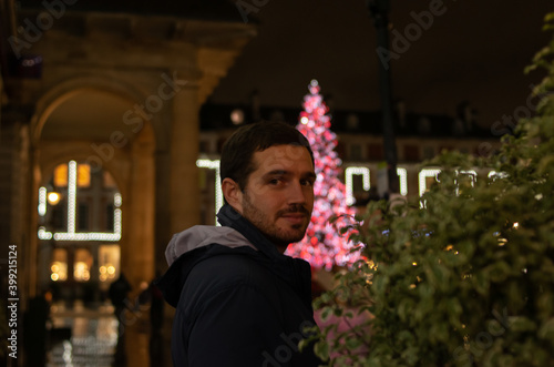 Obraz na płótnie Photo of a young and attractive man smiling next to a Christmas tree at night full of beautiful lights. Christmas decorations. ornaments w salonie